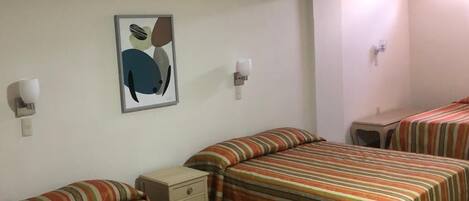 Habitación de hotel con tres camas, cada una con ropa de cama a rayas, una mesita de noche pequeña, una lámpara de pared y un cuadro enmarcado en la pared.