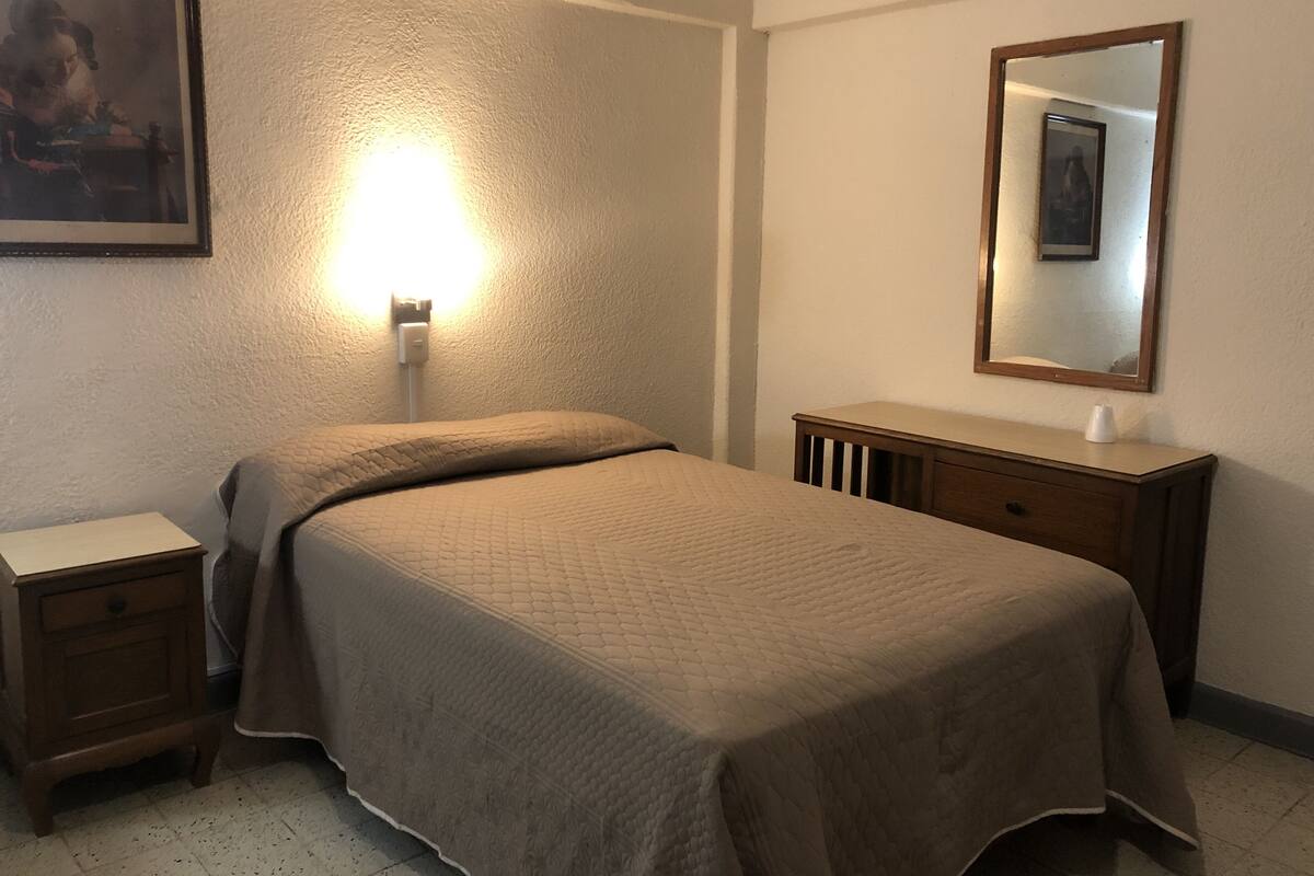 Quarto com cama, mesa de cabeceira, cômoda e luminária na parede.