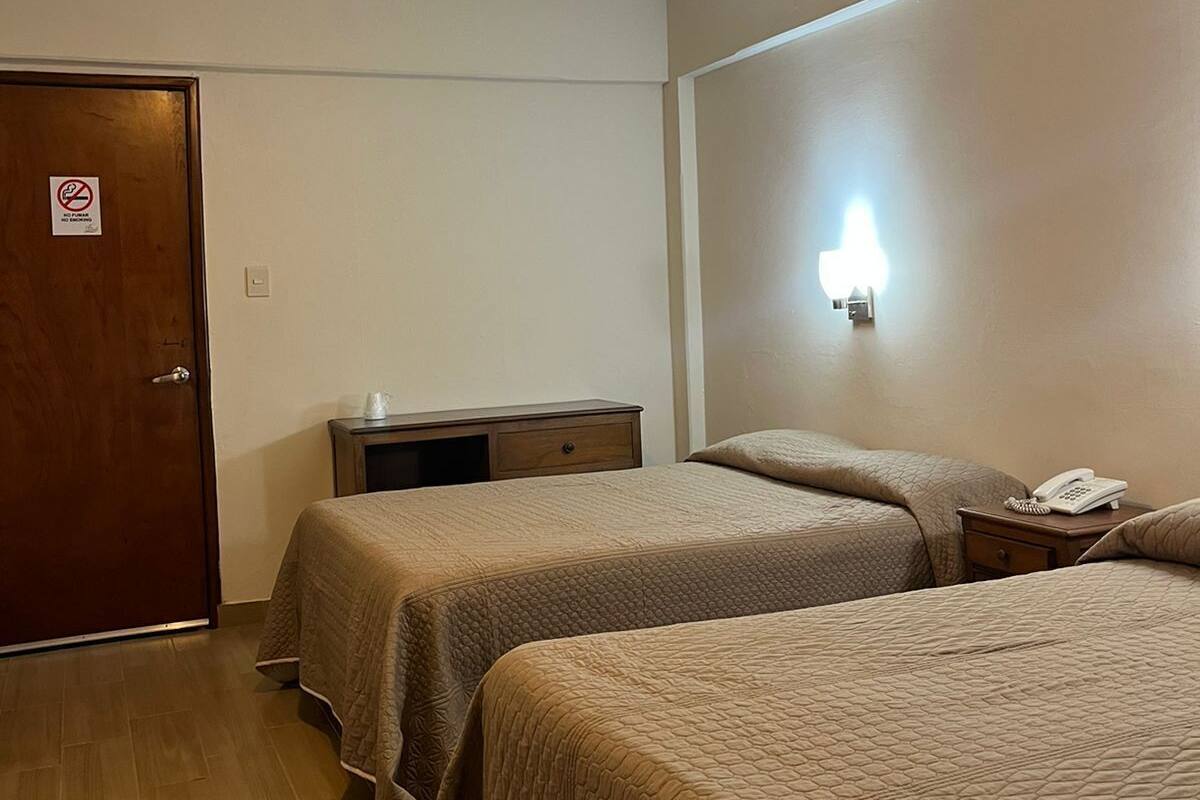 Quarto de hotel com duas camas, uma escrivaninha, um telefone na mesinha de cabeceira e uma porta com o aviso 'Não Fumar'.