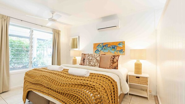 A hotel room with a bed, a ceiling fan, an air conditioning unit, a window with curtains, a painting, and a nightstand with a lamp.