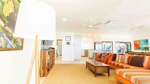 A spacious living room with a sofa, coffee table, and a TV. There is a kitchen in the background with a dining area.
