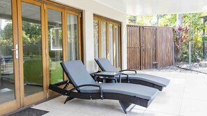 A covered outdoor seating area with two lounge chairs and a small table, adjacent to a sliding glass door leading to a garden.