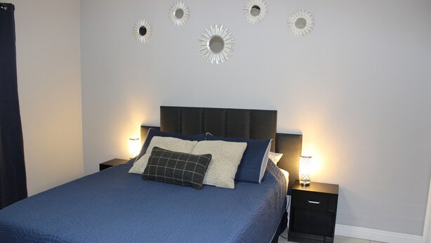 En pænt redt seng med et blåt sengetæppe og puder, med to sengeborde med lamper på hver side, i et rum med et enkelt, rent design.
