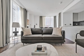 A modern living room with a grey sofa, a coffee table with books, and a view of the city through large windows.