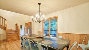 Un espace repas comprenant une table en bois, des chaises assorties, un lustre et une vue dégagée grâce à de grandes fenêtres.