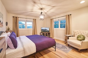 Quarto com uma cama, televisão, cadeira e duas janelas com cortinas.