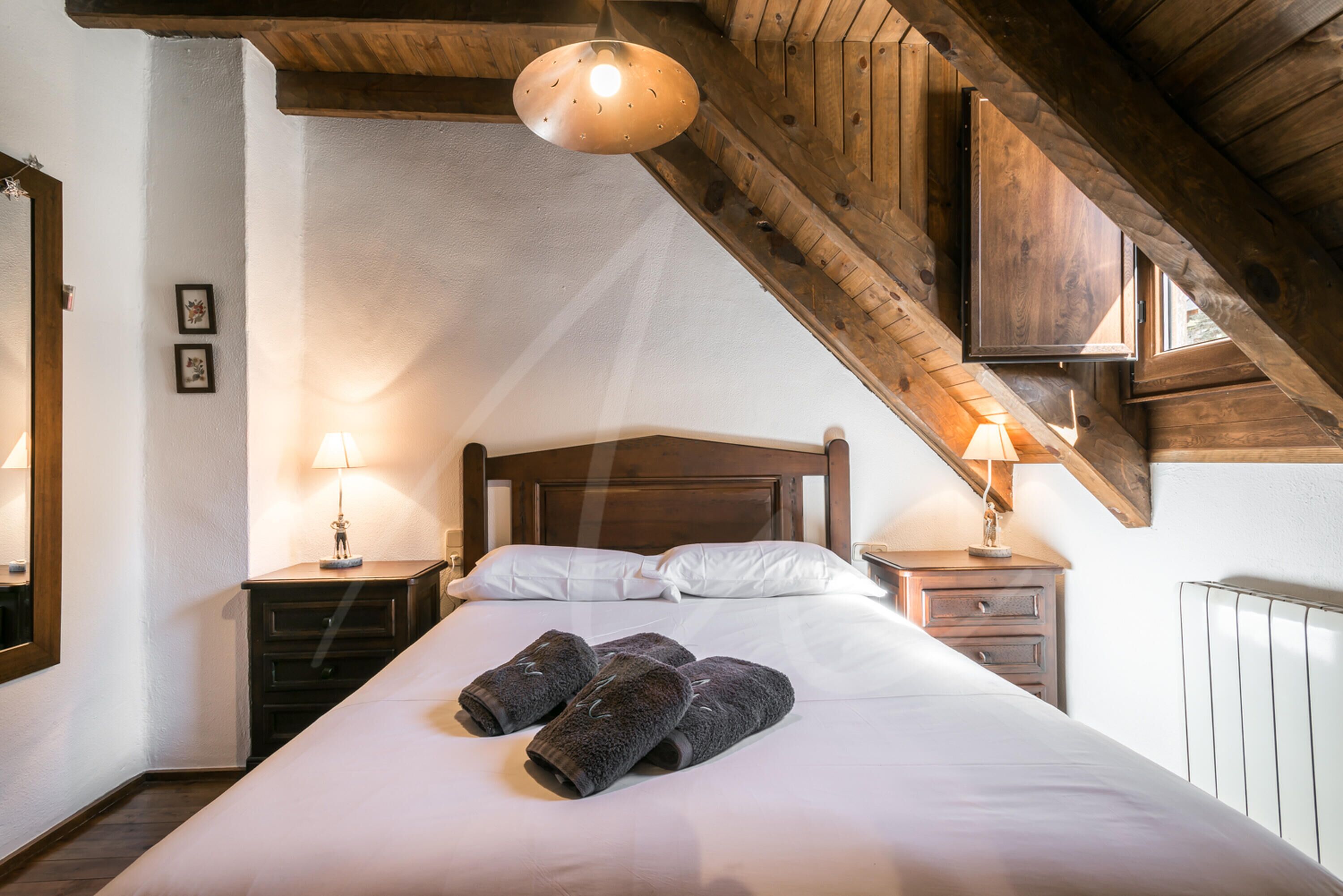 Une chambre avec un plafond en bois, un lit recouvert de draps blancs, deux tables de chevet avec des lampes et un radiateur.