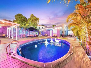 Une piscine avec une terrasse en bois, entourée d’espaces de détente en plein air et d’une zone couverte illuminée de guirlandes lumineuses.