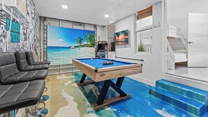 A room with a pool table, a sofa, a mural of a beach, and a staircase.