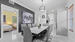 A modern dining area with a white table and grey chairs, a large painting on the wall, and a ceiling light fixture.