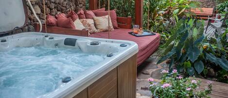A hot tub with a wooden deck, a wooden bench with cushions, and lush greenery.