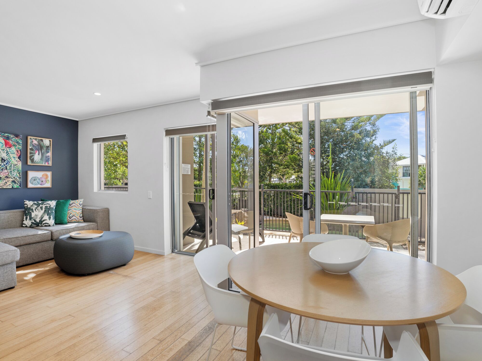 A modern living room with a grey sofa, a round wooden table, and a glass door leading to an outdoor patio.