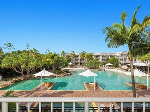 Zona de piscina de un resort con varias piscinas, palmeras y un edificio con balcones al fondo.