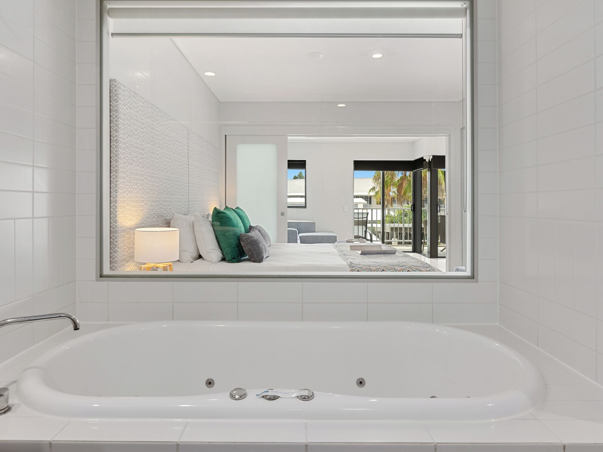 A modern bathroom with a large bathtub, a mirror reflecting a bedroom with a bed and pillows, and a view of a balcony with outdoor furniture.