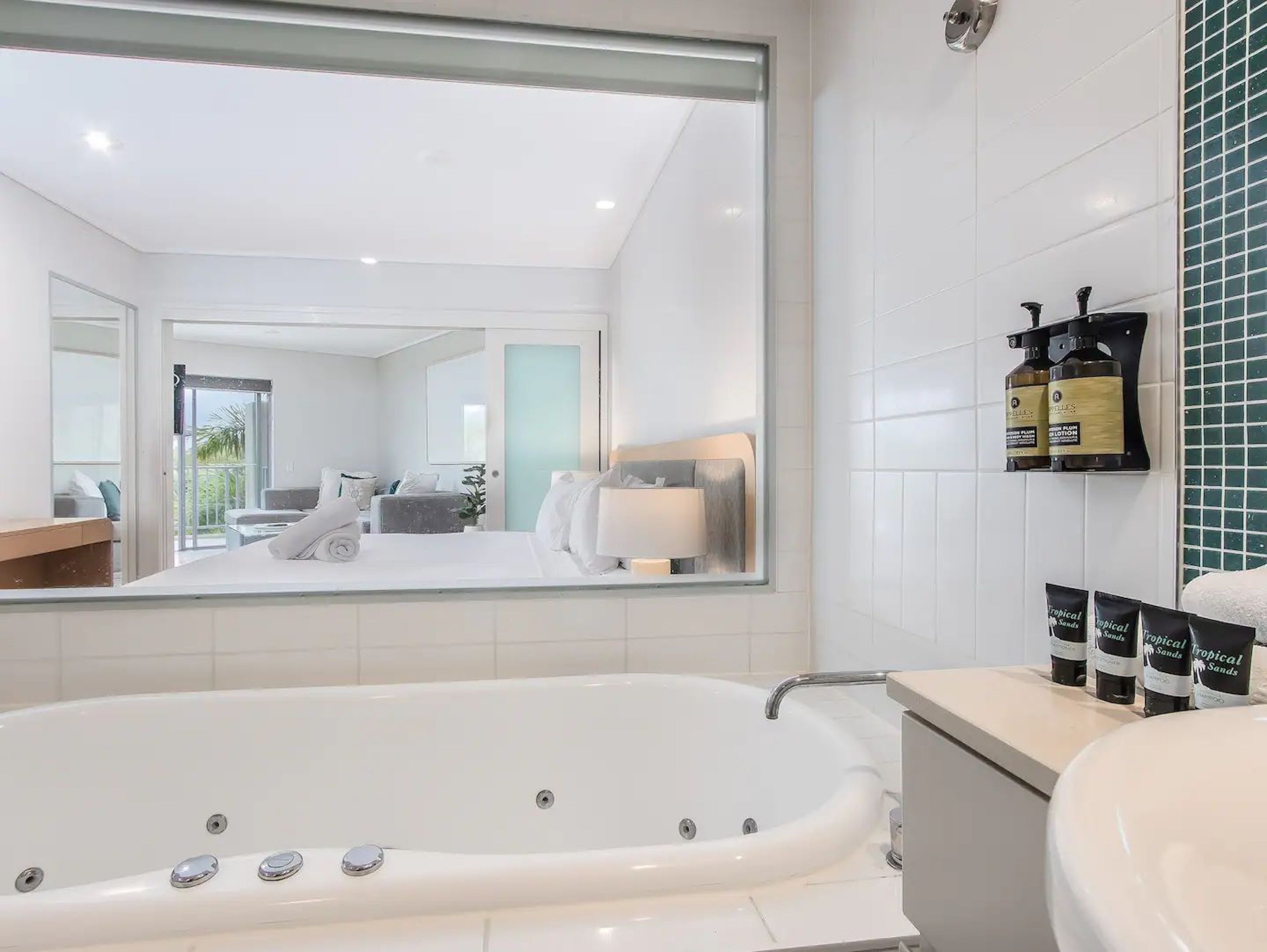 A modern bathroom with a large bathtub, a sink, and a mirror. There are toiletries on the counter and a view of the bedroom through the mirror.