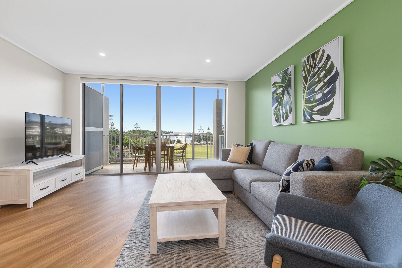 A modern living room with a large TV, a sofa, a coffee table, and a balcony with a view.
