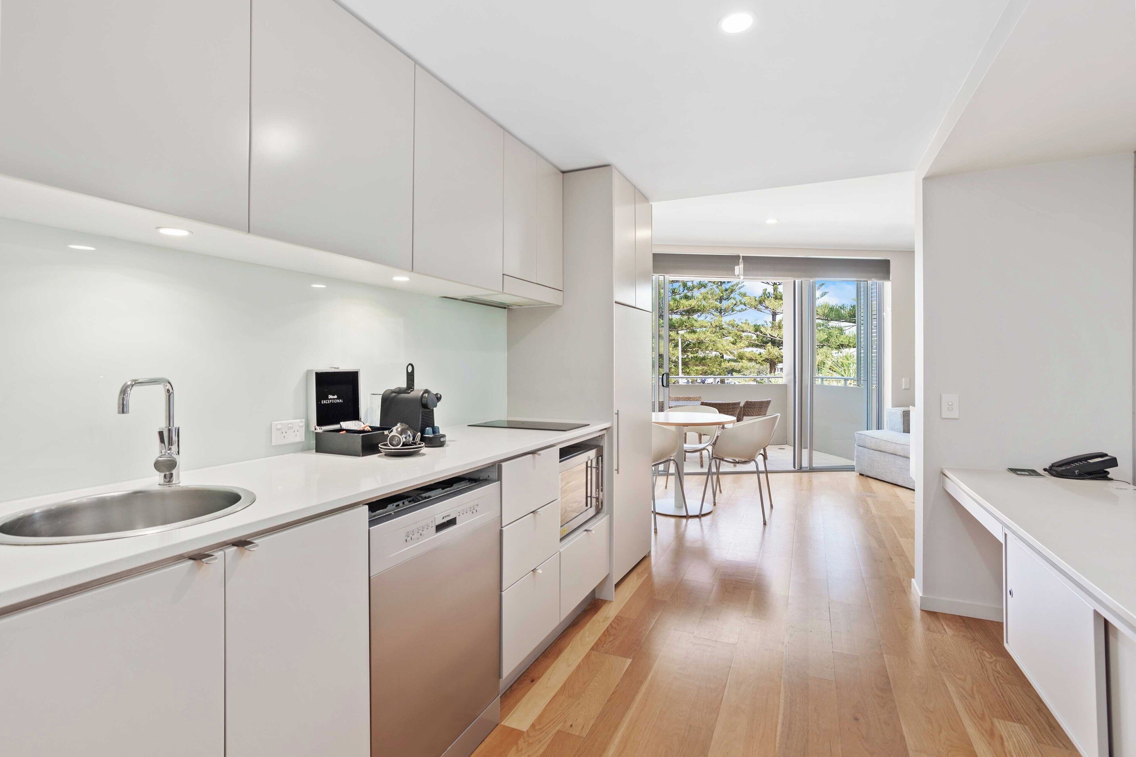 A modern kitchen with white cabinetry, a stainless steel sink, and a built-in dishwasher. There is a dining area with a table and chairs, and a view of the outdoors through large windows.