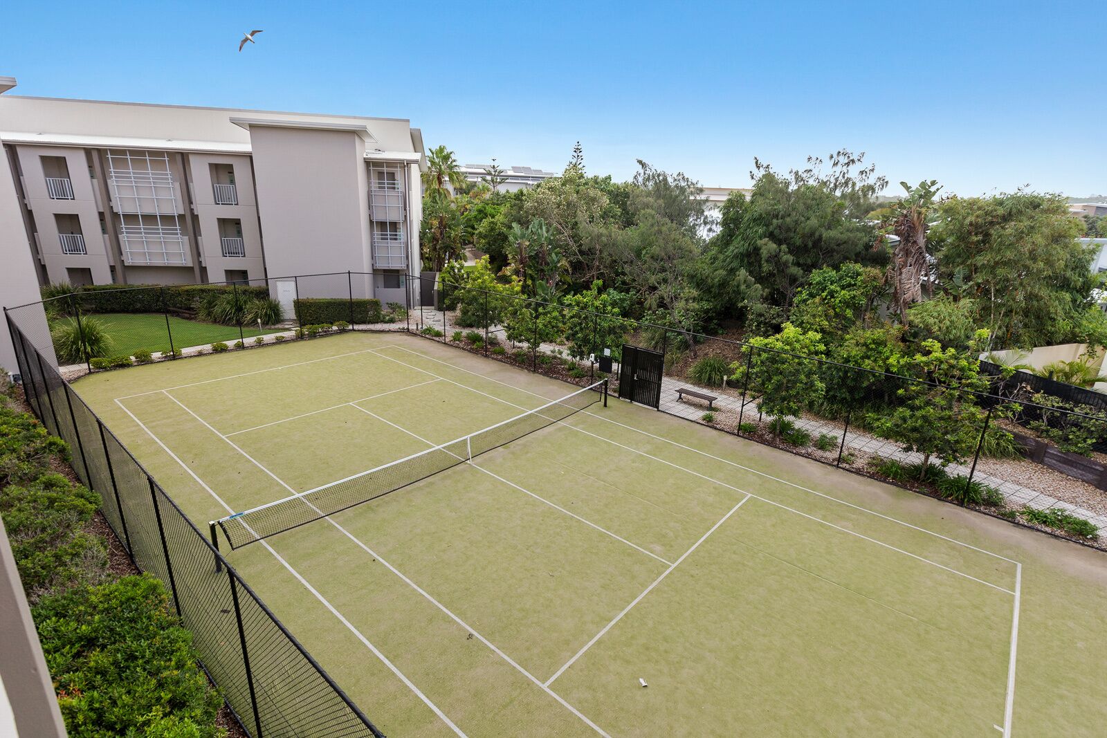 Una cancha de tenis con una red, rodeada por una cerca y follaje, con un edificio al fondo.