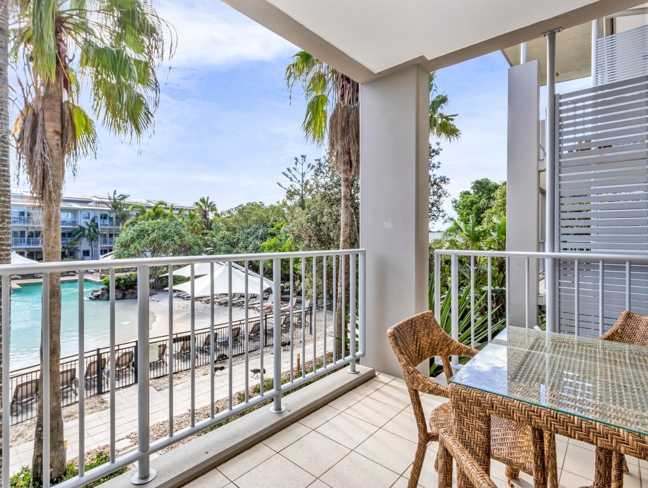A balcony with wicker furniture, a glass table, and a view of a pool and beach.