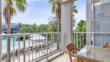 A balcony with wicker furniture, a glass table, and a view of a pool and beach.