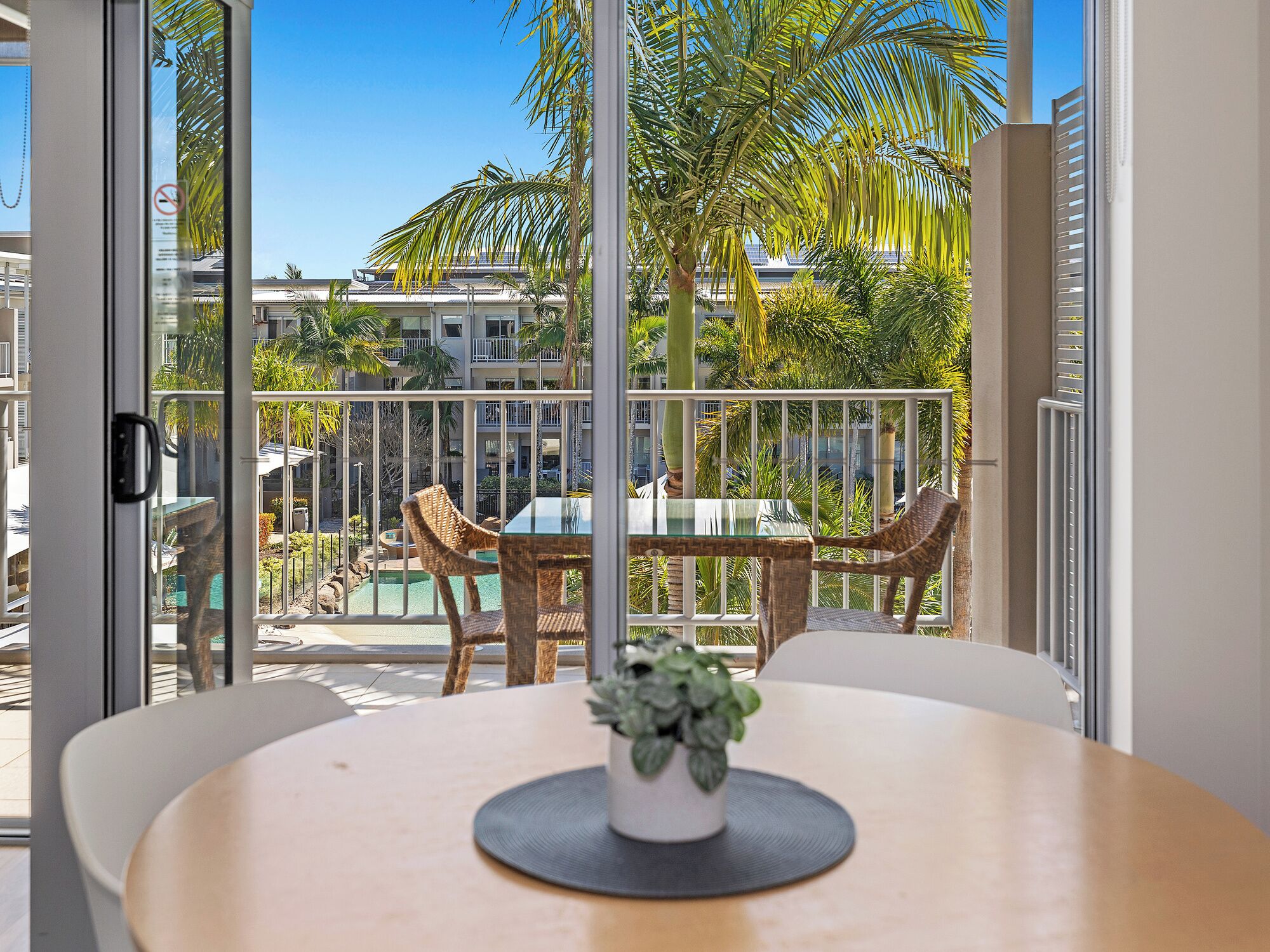 A balcony with a glass door, wicker chairs, a glass table, and a potted plant.