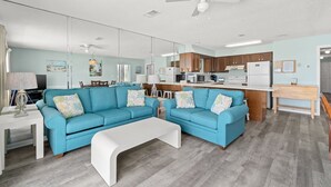 A modern living room with a blue sofa set, a white coffee table, and a kitchen area with wooden cabinets and a white refrigerator.
