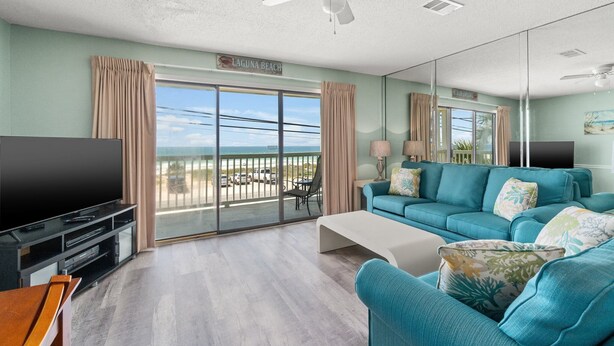 A living room with a large flat-screen TV, teal sofa, and a balcony with a view of the ocean.