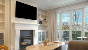 A modern living room with a fireplace, a flat-screen TV mounted above it, and a large window offering a view of the outdoors.