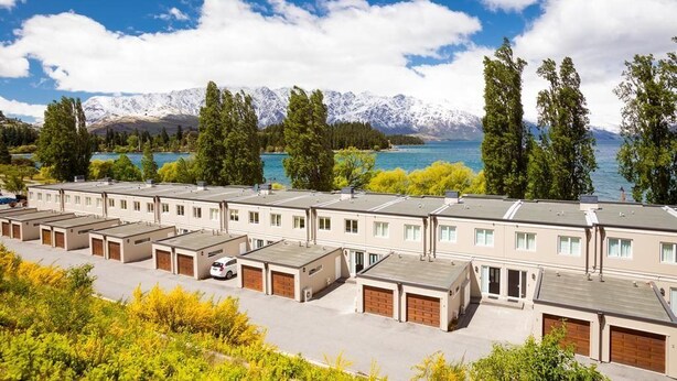 A row of modern, single-story buildings with attached garages, set against a backdrop of snow-capped mountains and a clear blue sky.