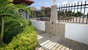 A gated entrance with a stone wall and metal fence, surrounded by well-maintained greenery and a clear sky. - Arena Condos Aruba 250 m to Eagle beach Aruba (Noord)