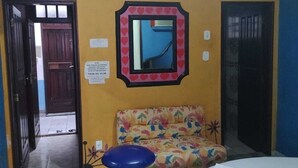 A room with a bed, a colorful sofa, a blue stool, a mirror with a heart border, and a door leading to another room. - Pousada Devaneios (Ananindeua)