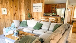 A cozy living room with a sectional sofa, wooden walls, and a kitchen area in the background. - Cozy Lakefront Bungalow w firepit/dock/kayaks (Plymouth)