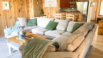 Un salon chaleureux avec un canapé d’angle, des murs en bois et un coin cuisine à l’arrière-plan.