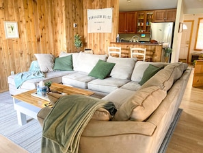 A cozy living room with a sectional sofa, wooden walls, and a kitchen area in the background.