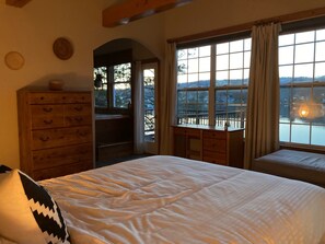 A bedroom with a bed, wooden dresser, desk, and large windows with a view of the outdoors.
