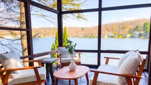 A wooden deck with a view of a lake and hills. There are two wooden chairs with white cushions and a small table with a vase. - Cozy Lakefront Bungalow w firepit/dock/kayaks (Plymouth)