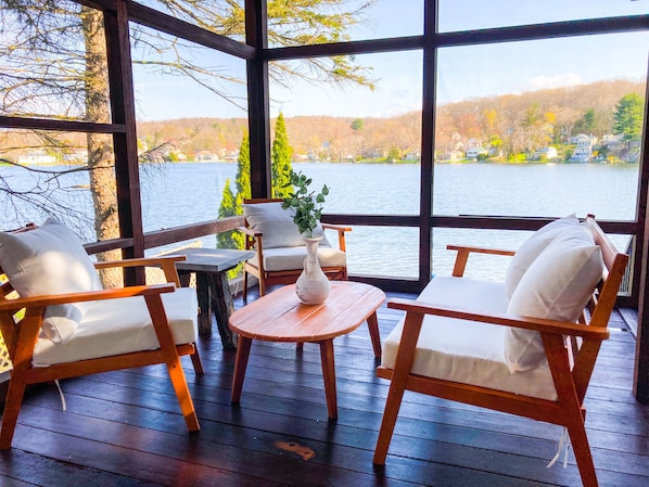 A wooden deck with a view of a lake and hills. There are two wooden chairs with white cushions and a small table with a vase.