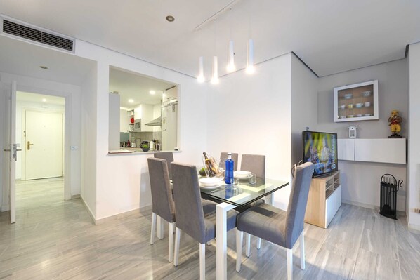 A modern dining area with a glass table, grey upholstered chairs, and a kitchen visible through an open doorway. - 2033 Beachfront Family Apartment on Golden Beach (Marbella)