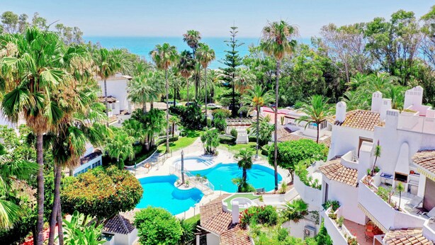 A resort with a central swimming pool surrounded by palm trees and a view of the sea.