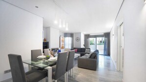 A modern living room with a glass dining table, grey upholstered chairs, a grey sofa, and a large window with curtains.