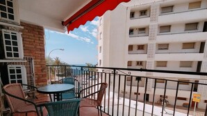 A balcony with a round table and chairs, a red awning, and a view of a building with barred windows. - Nerja Sun Apartment Bahía 17 (Nerja)