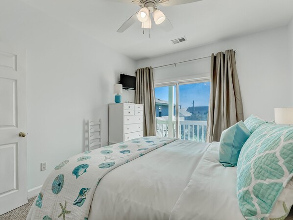 A bedroom with a bed, a dresser, a ceiling fan, a window with curtains, and a view of the outside.