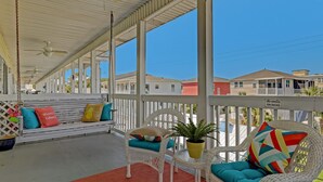 A covered porch with a white railing, a bench with colorful cushions, wicker chairs, and a small table with a plant. - South Beach Condo, Porch Swing with Ocean View (Tybee Island)