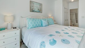 A bedroom with a bed, nightstands, a ceiling fan, a painting, and a bathroom visible through an open door. - South Beach Condo, Porch Swing with Ocean View (Tybee Island)