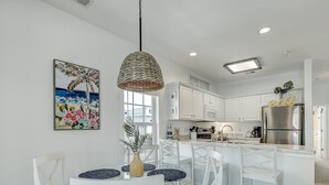 A modern kitchen with white cabinetry, a stainless steel refrigerator, and a dining area with white chairs and a glass table. - South Beach Condo, Porch Swing with Ocean View (Tybee Island)