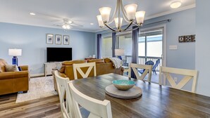 A dining area with a wooden table, white chairs, and a chandelier. There is a television and a window with curtains in the background. - South End Tybee Getaway, Short Walk to Beach (Tybee Island)