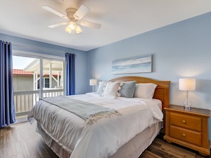 Une chambre à coucher avec un lit, une table de chevet, un ventilateur de plafond, une fenêtre avec des rideaux et un tableau au mur.