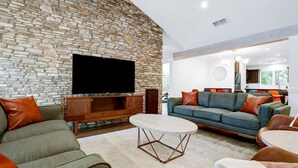 A living room with a stone wall, a flat-screen TV, a sofa, a coffee table, and a dining area with chairs. - Pool~Hottub~Yoga Gym~Game Room~Fire Pit (Austin)