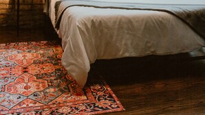 Ein Schlafzimmer mit Backsteinwand, großem Fenster mit Vorhängen, Teppich mit Muster und einem Bett mit vielen Kissen.