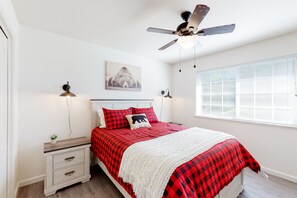 A bedroom with a bed, nightstands, a ceiling fan, a window with blinds, and a wall-mounted lamp.